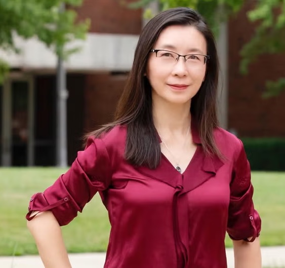 A woman in a collared shirt, hand son hips, poses in a courtyard